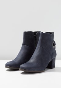 Botas de tobillo azul marino de cuero liso con cremallera lateral, puntera redondeada y un acento circular cerca del tobillo. Tacón grueso.