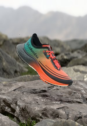 Trailrunning-Schuh mit grün-orangem-schwarzem Farbverlauf, Columbia-Logo und roten Schnürsenkeln, in der Luft über felsigem Gelände fotografiert.