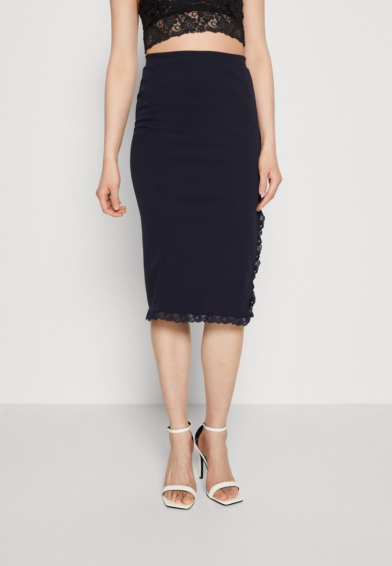 Femme portant une jupe crayon bleu marine cintrée longueur genou avec bordure en dentelle et des sandales blanches à talons hauts avec bride autour de la cheville, debout sur un fond uni.