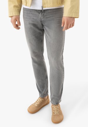 Mężczyzna w jasnoszarych dżinsach, beżowych trampkach i żółtej kurtce stoi na białym tle.