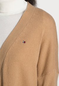 Cárdigan beige hecho de material de punto, con un profundo escote en V y un pequeño logo bordado con acentos en rojo, blanco y azul.