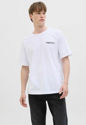 Joven con camiseta blanca con texto negro en el pecho izquierdo, combinado con jeans negros, posando contra un fondo liso y claro.