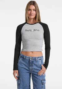 Gray cropped long-sleeve shirt with black raglan sleeves, featuring a silver Guess Jeans logo on the front. Casual fit and smooth texture.
