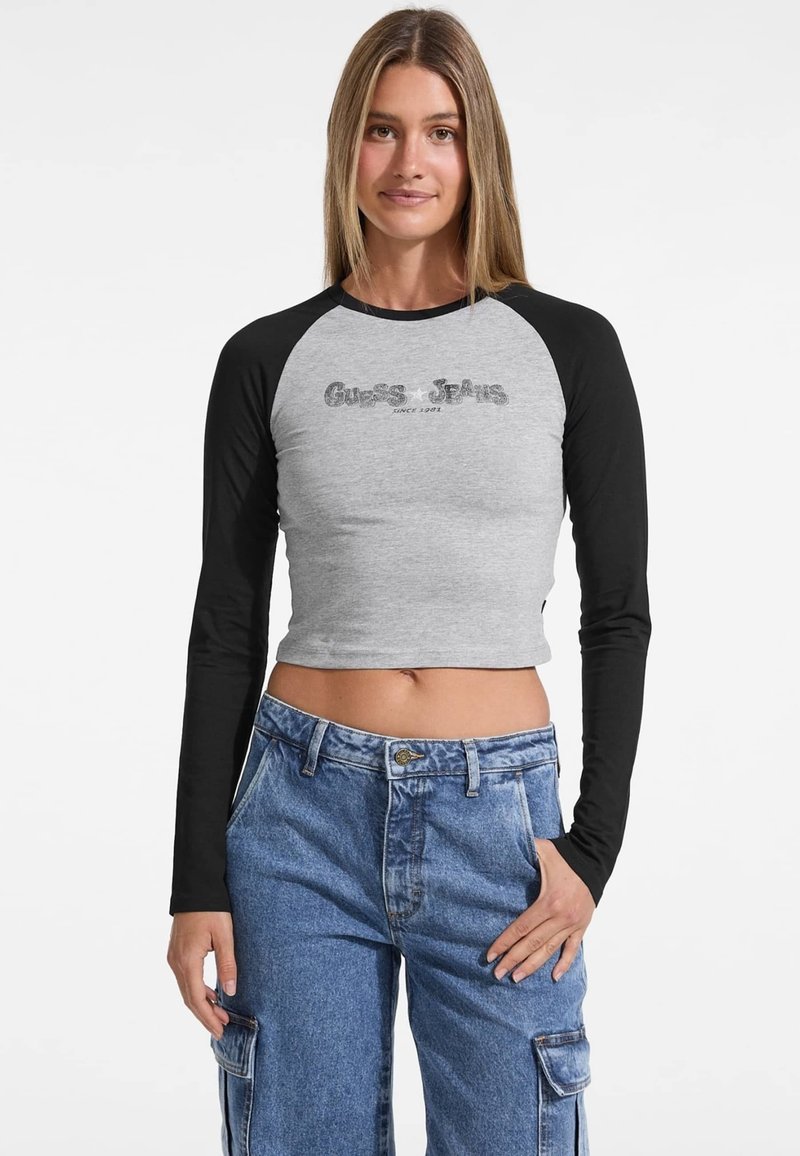 Siva skrajšana dolga majica z dolgimi rokavi z črnimi raglan rokavi, z usnjenim logotipom Guess Jeans spredaj. Udobna prileganje in gladka tekstura.