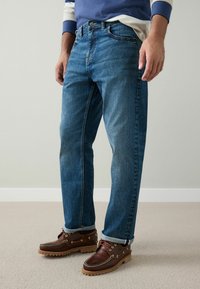 Niebieskie dżinsy denimowe o prostym kroju nogawki, wyposażone w klasyczny design z pięcioma kieszeniami. Noszone z brązowymi skórzanymi butami i podwiniętymi mankietami.