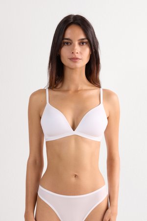 Mujer con cabello largo y oscuro, vestida con un sujetador blanco y bragas a juego, de pie frente a un fondo claro liso, mirando hacia adelante.