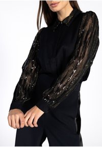 Vrouw in zwarte blouse met doorzichtige, pailletten lange mouwen en bijpassende zwarte hoge taille broek, staand tegen een witte achtergrond.