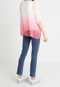 Transparante blouse met een verloop van wit naar roze, ronde halslijn, korte mouwen, gecombineerd met blauwe jeans en witte sneakers.