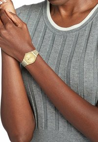 Gold-tone wristwatch with a round face, mesh band, and light texture, worn on a wrist against a gray ribbed knit top.