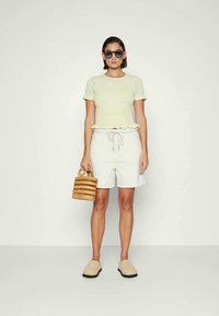 Camisola de malha amarelo claro com mangas curtas e bainha em franzido, combinada com calções brancos, sapatos bejes de slip-on e uma bolsa tecida bege.