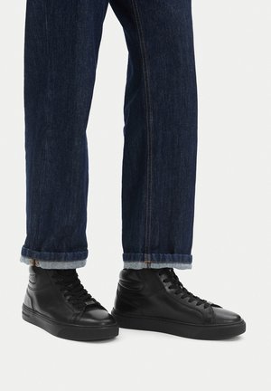 Osoba ubrana w podwinięte ciemnoniebieskie dżinsy i czarne skórzane sneakersy za kostkę z czarnymi sznurowadłami na jednolitym tle.