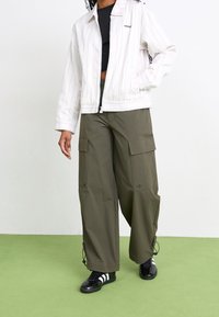 Bílá pruhovaná bunda na zip, olivově zelené cargo kalhoty s kapsami a černé sportovní boty s bílými pruhy. Na pozadí zelená podlaha.