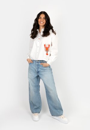 Smiling woman stands with hands in pockets wearing white shirt with red lobster graphic, light blue wide-leg jeans, and white sneakers.