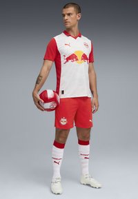 Equipación de fútbol roja y blanca con una camiseta que lleva un logo de toro rojo, pantalones cortos rojos, calcetas blancas con acentos rojos y un balón de fútbol con un diseño en rojo y blanco.