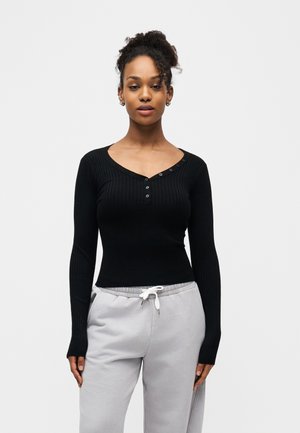 Göndör hajú nő fekete, bordázott, gombos hosszú ujjú ingben és világosszürke, fehér zsinórral ellátott melegítőnadrágban, fehér háttér előtt áll.