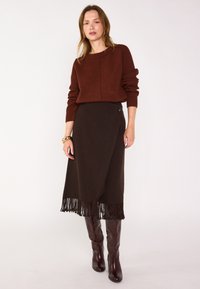 Maglione marrone a coste con scollatura rotonda, abbinato a una gonna avvolgente marrone scuro con dettagli in frange e bottoni. Stivali marroni al ginocchio.