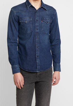 Homme portant une chemise en denim bleu foncé cintrée avec des poches à rabat et un jean noir sur un fond clair uni.