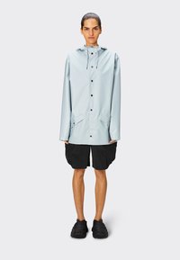 Chaqueta impermeable azul claro con capucha, botones de presión en la parte frontal y dos bolsillos laterales, combinada con pantalones cortos negros texturizados y zapatos negros.