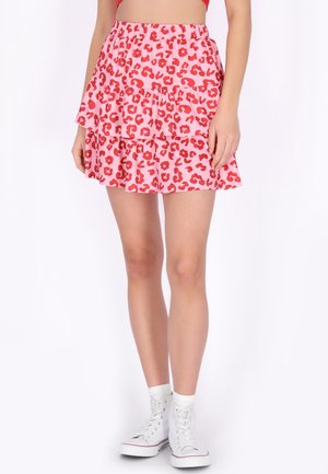 Frau trägt einen rosa gestuften Minirock mit roten Leopardenflecken und weißen High-Top-Sneakern, steht vor einem einfarbigen Hintergrund.