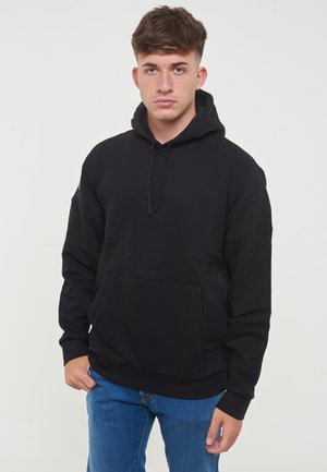 Felpa nera con tasca a canguro, cappuccio con cordino e cuciture rinforzate. Indossata con jeans blu, design minimalista e texture morbida.