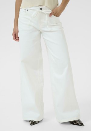 Kvinde iført hvide vidde bukser med høj talje og bindebælte, hånd i lommen, og synlige zebra-print spidse sko.