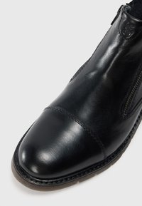 Botín negro de cuero con puntera, cremallera lateral y suela resistente sobre un fondo claro.