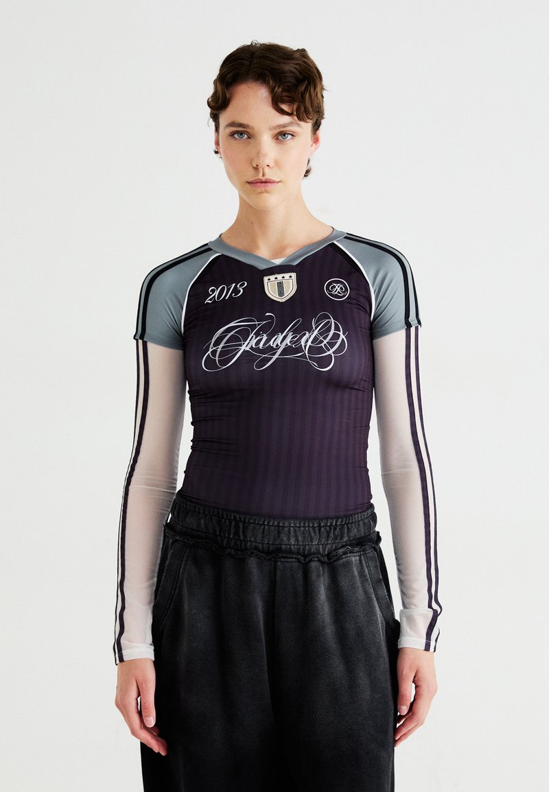Cămașă sport cu mânecă lungă, de culoare mov cu dungi verticale, având accente gri, decupaj în formă de V și detalii grafice, asociată cu pantaloni casual închisi la culoare.