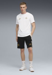 Camiseta blanca de manga corta con detalles dorados, combinada con pantalones cortos negros que presentan logotipos dorados y una franja lateral con un patrón. Terminada con zapatillas blancas.