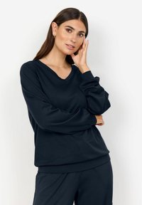 Hosszú barna hajú nő sötét navy V-nyakú pulóverben és hozzá illő nadrágban pózol, egyik kezével az arcához érve, fehér háttér előtt.