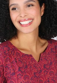 Das rot und lila gemusterte Oberteil zeichnet sich durch ein florales Design, einen runden Halsausschnitt und einen weichen Stoff aus. Der Hautton wirkt ebenmäßig mit natürlichem Makeup.