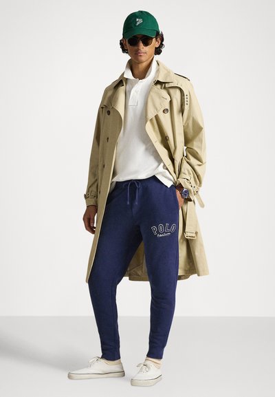 Béžový trenčkot cez bielu polokošeľu, v kombinácii s tmavomodrými teplákmi s logom "POLO". Zelená čiapka a čierne slnečné okuliare, biele tenisky.