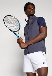 Marinblå vadderad väst med zip framtill, vit logotyp, i kombination med en marinblå polo och vita shorts. Håller en tennisracket, mot en enhetlig bakgrund.