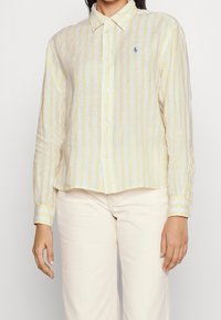 Linen shirt featuring vertical yellow and white stripes, button-down collar, and small blue logo. Long sleeves with buttoned cuffs.