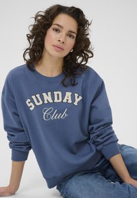 Žena s kovrčavom kosom u plavom dukserici s natpisom "SUNDAY Club" i plavim trapericama, sjedi s jednom rukom oslonjenom na pod.