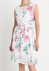 Blommig klänning gjord av lätt tyg, med ärmlös design, rund halsringning och en knytbälte. Färgerna inkluderar vitt, rosa och teal.