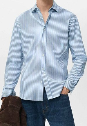 Mężczyzna w niebiesko-białej koszuli w pionowe paski, niebieskich dżinsach, trzyma brązową kurtkę w lewej ręce, prawą rękę ma w kieszeni.