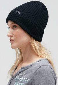 Black ribbed knit beanie with a folded cuff, featuring a small tag. Accompanied by a striped gray top, showcasing a casual design.