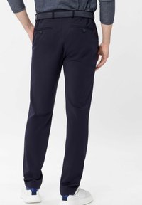 Homme portant un pantalon ajusté bleu marine, une chemise à motifs rentrée à l'intérieur, et des baskets blanches, se tenant contre un fond uni.
