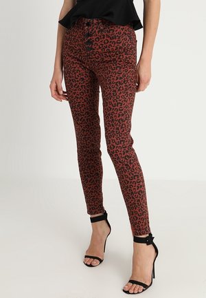 Nainen pukeutunut ruosteenvärisiin leopardi-kuosisiin kapeisiin housuihin, joissa on mustat nappisulkeet, yhdistettynä mustiin hihkaisiin korkokenkäsandaaleihin ja mustaan toppiin.