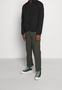 Schwarze Bomberjacke mit Reißverschluss vorne, kombiniert mit olivgrünen Hosen und grünen High-Top-Sneakers mit weißen Akzenten.