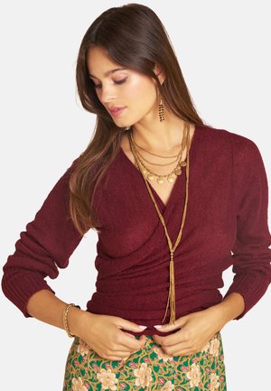 Frau mit langen braunen Haaren trägt einen bordeauxfarbenen gestrickten Wickelpullover, geschichtete Goldketten, goldene Ohrringe und einen Rock mit Blumenmuster und richtet ihren Pullover.