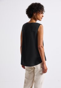 Blusa nera senza maniche con una texture liscia, orlo arrotondato e dettagli discreti sulla parte posteriore; abbinata a pantaloni beige chiaro a vestibilità comoda.