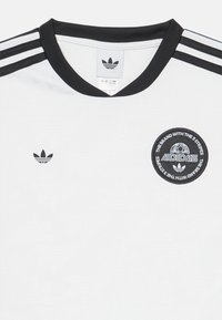 Camiseta blanca de Adidas con cuello de canalé negro, tres franjas negras en los hombros y un logo circular en el pecho.
