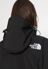 Personne aux cheveux bruns portant une veste noire avec une grande capuche et le logo blanc The North Face dans le haut du dos.