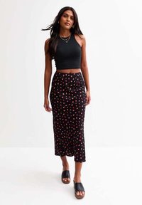 Débardeur noir court associé à une longue jupe noire ornée d'un motif floral rouge, complété par des chaussures noires à enfiler.