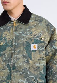 Giacca con motivo mimetico in una combinazione di colori verde e marrone, colletto in velluto a coste, chiusura con zip e un'etichetta con marchio sulla tasca del petto.