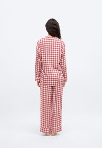 Γυναίκα όρθια, ξυπόλυτη, φοράει σετ πιτζάμες με κόκκινο και λευκό καρό και σχέδια καρδιών, στραμμένη προς τα πίσω σε απλό λευκό φόντο.