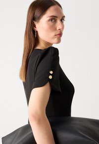 Juodas prigludęs suknelė su pūstomis rankovėmis, aukso mygtukų akcentais ir lygiu paviršiumi. Modelis laiko didelę juodą rankinę.