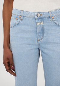 Jean bleu clair porté avec un top blanc, montrant la taille, le bouton, les poches et une main aux ongles vernis reposant sur le côté.