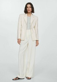 Svetlo obarvan tailored blazer z teksturiranim zaključkom, v kombinaciji s sivo majico in ohlapnimi belimi hlačami. Črne natikače dopolnjujejo celotno kombinacijo.